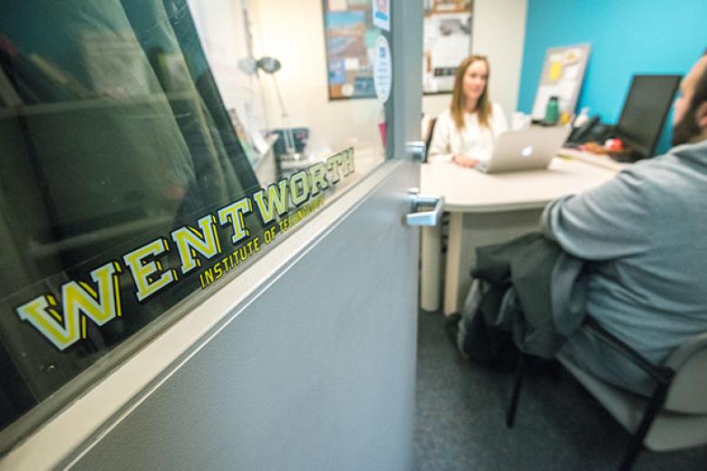 In the foreground, the focus is on a Wentworth decal on an office door. In the background a student sits across from a female advisor at her desk.