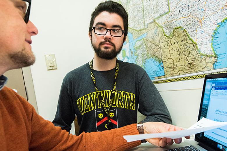 A male student looks on as an advisor talks to him in front of his laptop.