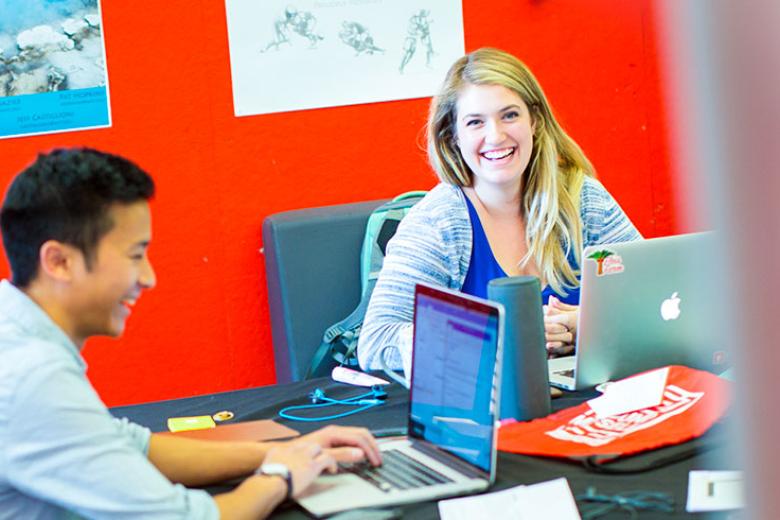 A female student smiles at the camera while a male student laughs as he looks at his laptop. 