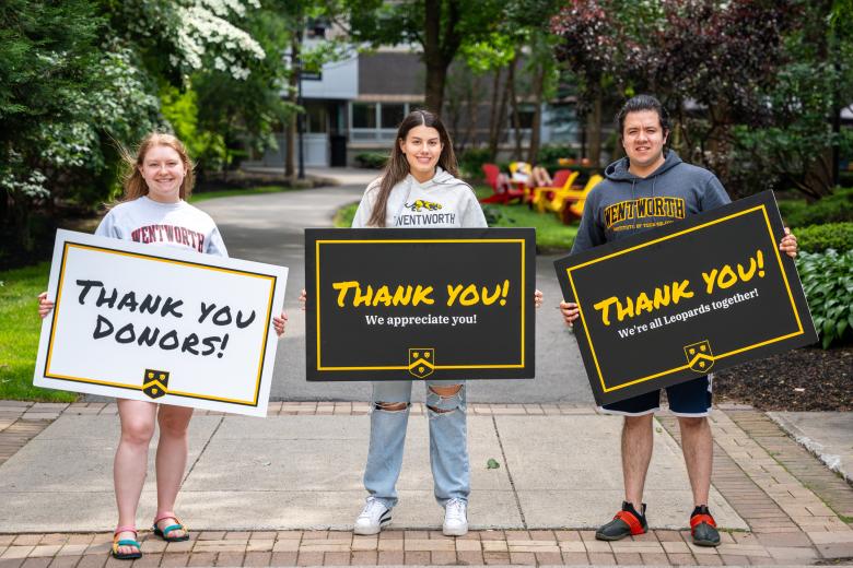 Three students wearing Wentworth sweatshirts hold up signs which say "Thank You Donors" on campus