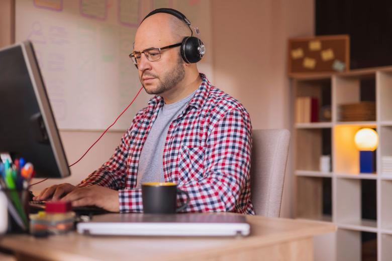 A man sits at home on his computer with headphones on while looking at the display