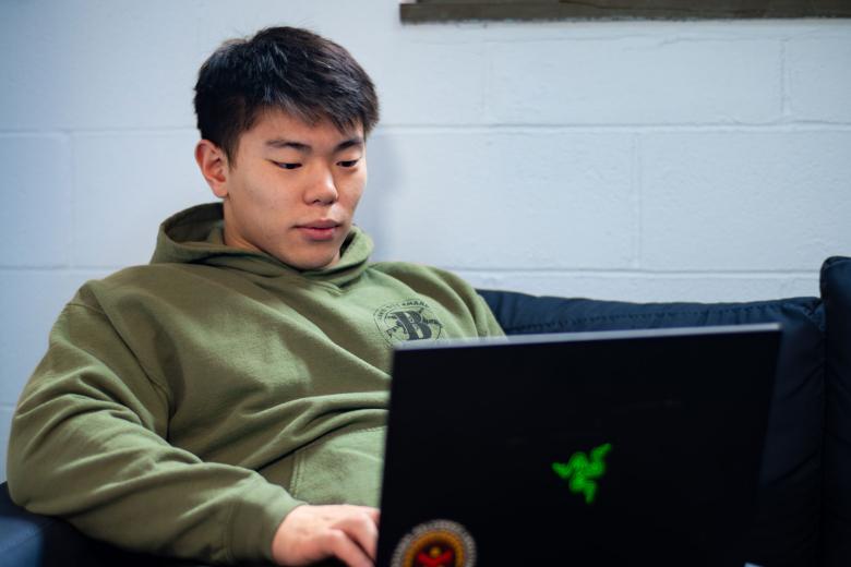 A student wearing a military hoodie studies at a laptop on a couch