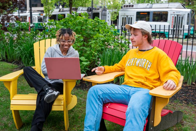 A Black female student and a white male wearing a Wentworth shirt sit together on the quad to study looking at a laptop. The Green Line T is in the background.